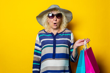 Old woman with surprised face in hat and glasses holds shopping bags with purchases on yellow background