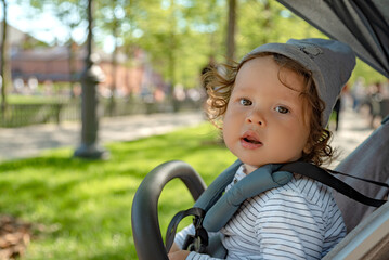 Close up portrait of an adorable toddler sitting in a pram and looking at the camera on the background of the green public park.