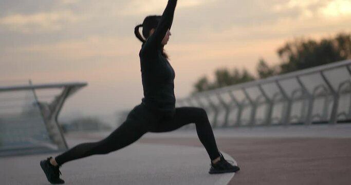 Fit female athlete lunging near running path. Side view of slim sportswoman with ponytail doing lunges and stretching legs near path during fitness training at sunset on embankment