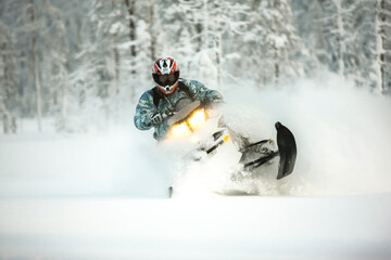 Fototapeta na wymiar The rider in gear with a helmet makes a sharp turn on a snowmobile on a deep snow surface on a background of snowy landscaping nature and winter forest.