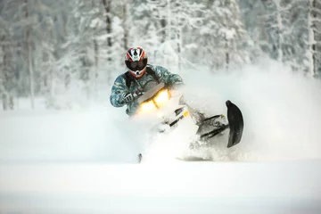 Fotobehang The rider in gear with a helmet makes a sharp turn on a snowmobile on a deep snow surface on a background of snowy landscaping nature and winter forest. © igor tsarev 