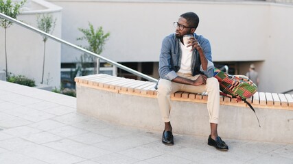 Young handsome African American man sitting with backpack on bench in city. Stylish guy is enjoying time and drinking coffee.