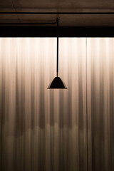Isolated metal pendant light hanging from the ceiling with sheer ripple curtain hanging in the background, lit at night 