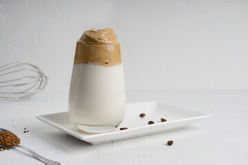 Glass of homemade dalgona drink made by whipping instant coffee powder, sugar and hot water with addition of cold milk on white plate with kitchen utensil on wooden background. Image with copy space