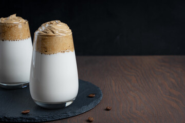 Glass of homemade dalgona drink made by whipping instant coffee powder, sugar and hot water with addition of cold milk on stony tray on dark wooden background at kitchen. Image with copy space