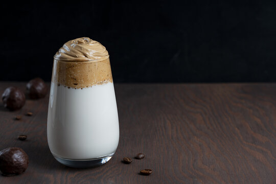 Glass of homemade dalgona drink made by whipping instant coffee powder, sugar and hot water with addition of cold milk with chocolate candies on dark wooden background. Image with copy space