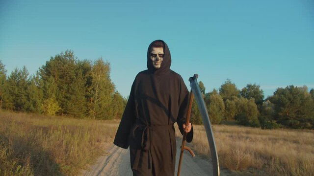 Creepy angel of death, grim reaper walking on dirt road at twilight, frightening with rustic scythe, expressing fear and ruthlessness in autumn nature while looking for victim on halloween.