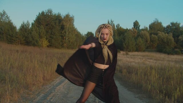 Spooky bloodthirsty female vampire with dreadlocks and bloody face hypnotizing, elegantly spinning in black cloak fluttering in wind on dirt road in rays of setting sun, expressing dangerous charm.