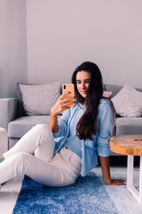 Obraz premium European fit brunette fashion blogger woman sits on floor in living room near sofa, wearing white jeans and blue sweather, and take photo selfie in mirror 