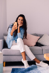 European fit brunette fashion blogger woman sits on floor in living room near sofa, wearing white jeans and blue sweather, and take photo selfie in mirror     