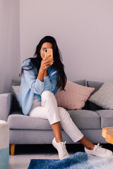 Obraz premium European fit brunette fashion blogger woman sits on floor in living room near sofa, wearing white jeans and blue sweather, and take photo selfie in mirror 