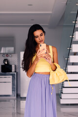 Fototapeta na wymiar Stylish caucasian fit pretty long hair woman fashion blogger wearing yellow top and purple pants, holding bright bag, make selfie take photo on mobile phone camera in mirror in kitchen at home. 