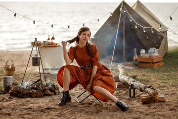 A girl in a brown dress sits near a campfire in front of a tent.