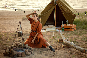 Beautiful girl in brown next to a campfire on the background of a lake.