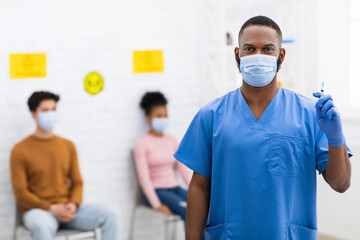 Fototapeta na wymiar Black Doctor In Face Mask Holding Syringe Standing In Hospital