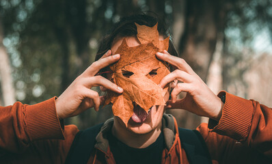 the guy covers his face with leaves and fools around. a man holds leaves with holes for eyes in front of his face. cheerful guy in the autumn forest. young man posing in front of the camera