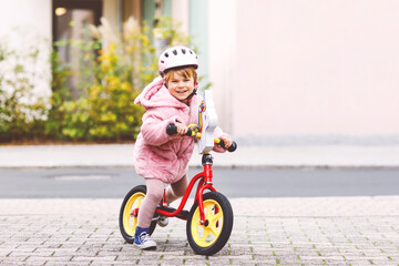 Fototapeta premium Cute little toddler girl with helmet riding on run balance bike to daycare, playschool or kindergarden. Happy child having fun with learning on learner bicycle. Active kid on cold autumn day outdoors.