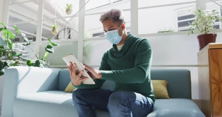 Mixed race man wearing face mask wiping his digital tablet with a tissue - Powered by Adobe