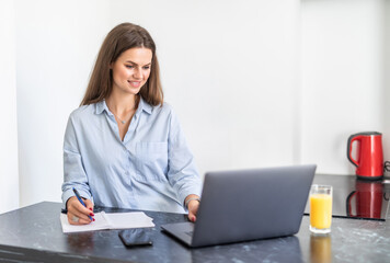 Young woman working on laptop writing in a notebook at home in the kitchen