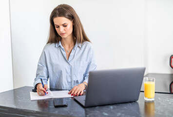 Young woman working on laptop at home on kitchen. writing on notepad. Business woman thinking about something important. Woman trying to find a job.