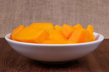 Pieces of pumpkin in the bowl at the wooden table and sackcloth background.