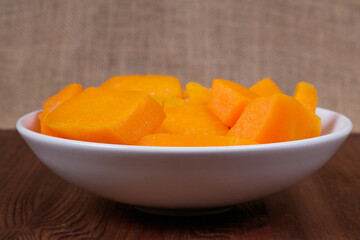 Pieces of pumpkin in the bowl at the wooden table and sackcloth background.
