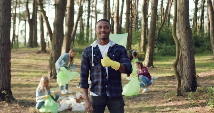 Likable contented active african american standing in front of camera near his friends-members of community of nature lovers which collecting into plastic bags rubbish from forest territory