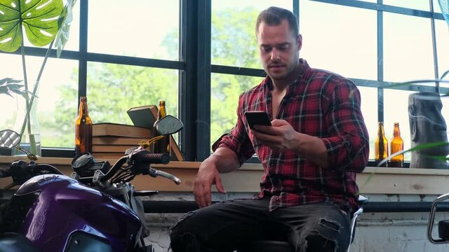 Hipster with smartphone relaxing in comfortable hall with plants and motorcycle around.