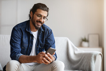 Handsome young indian guy sitting with smartphone at home, messaging with friends