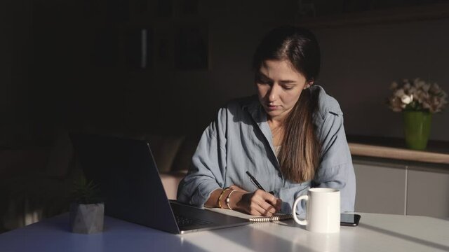 A young woman sits at a table and works on a laptop online from home