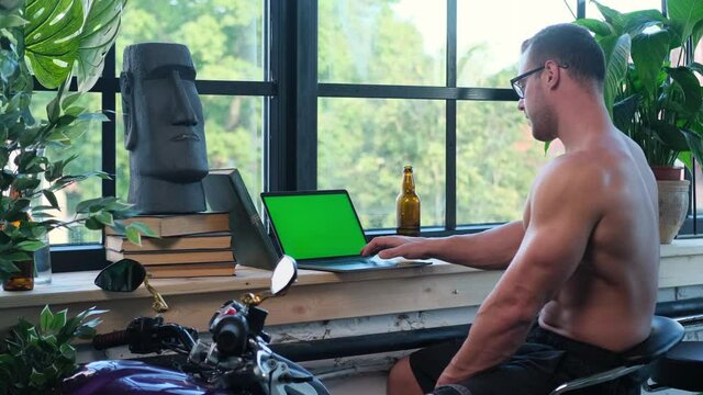 Shirtless sport guy with glasses and notebook relaxing in hall with interesting design with motorbike and plants.