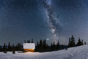 Fantastic winter landscape with wooden house in snowy mountains. Starry sky with Milky Way and snow covered hut. Christmas holiday and winter vacations concept