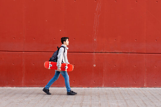 Side View Of Teenage Boy Walking On Sidewalk With Skateboard