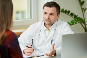 A caucasian doctor in a white lab coat is sitting at a desk and describing the treatment to a...