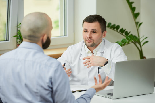 A Caucasian Doctor In A White Lab Coat Is Sitting At A Desk And Arguing About The Treatment Of A Bald Male Patient In A Hospital. A Man With A Beard At An Appointment In A Doctor's Office.