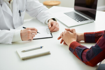 A photo of the folded hands of a patient who is gesturing near a doctor during an appointment in a hospital. A notebook with a pen, a clipboard, a laptop are lying on a white desk in a doctor's office