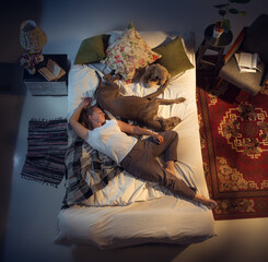 Obraz premium Portrait of a woman, female breeder sleeping in the bed with her dog at home. Top view. Dressed housekeeper sleeping after tiring work day. Holding his pet close to. Job, work, pets love concept.