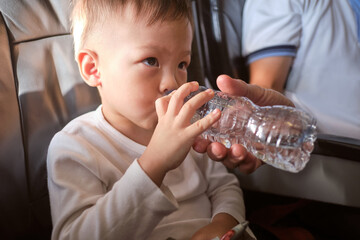 Cute little Asian 3 years old toddler boy child drinking water from bottle during flight on airplane. Little kid feeling earache on airplane