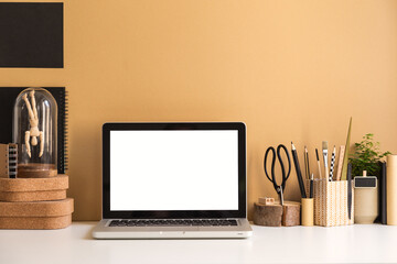 Desk with laptop mockup, stationery, and wood supplies near brown, beige wall.
