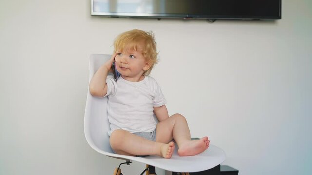 adorable blue eyed little son wearing clean white t-shirt and grey bloomers sits on chair holding modern cell phone near ear in light room