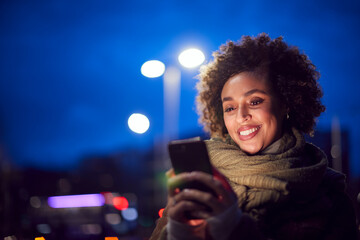 Woman On City Street At Night Ordering Taxi Using Mobile Phone App