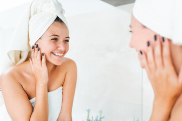 Young attractive woman with white towel on head and body with reflection in mirror in bathroom. Female smile and look at herself