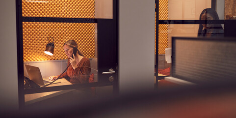 Businesswomen Working Late In Individual Office Cubicles Using Laptop And Digital Tablet