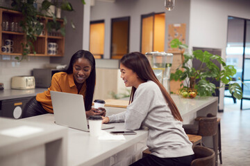 Two Young Businesswomen Working On Laptop In Kitchen Area Of Modern Office