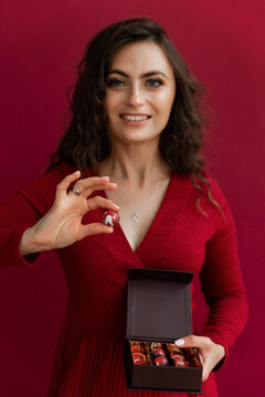 Pretty emotional woman in red dress posing with little box of bonbons and happy smiling in studio. Beautiful female holding one round candy. Conept of Christmas and New Year holidays