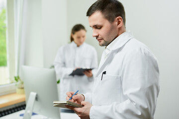 Fototapeta premium A caucasian doctor with a notebook and a pen records the patient’s medical history in a hospital. A female therapist is doing notes in a clipboard near an all in one computer in a clinic.