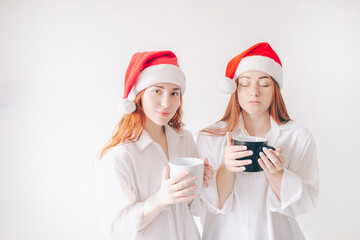 Two red-haired sisters stand isolated on a white background in spacious oversized shirts. Merry Christmas and happy new year. Two women in red santa claus hats drink tea or coffee, look at the camer .