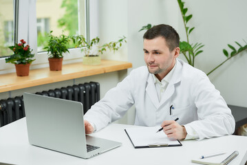 Fototapeta premium A happy caucasian doctor in a white lab coat is listening to a patient on an online meeting on a computer in a hospital. A therapist is sitting at a desk with a laptop in a doctor's office.