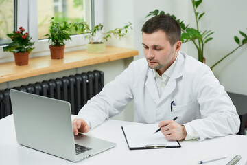 A serious caucasian doctor in a white lab coat is listening to a patient on an online meeting on a computer in a hospital. A therapist is sitting at a desk with a laptop in a doctor's office.