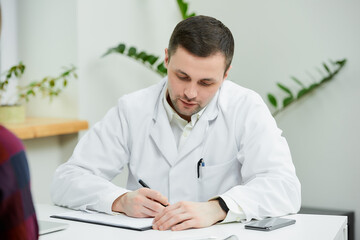 A portrait of a doctor in a white lab coat who is sitting at a desk and writing a diagnosis to a...